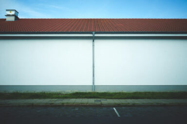 Experience the simplicity and elegance of this minimalistic digital photo featuring a white building wall with a red roof, surrounded by green grass and a small parking area in the front. The sunlit scene casts harsh shadows, creating a dramatic effect against the clear blue sky. This oddly empty image is strictly composed and simplistic, allowing you to appreciate the beauty of the architecture and natural surroundings.  Get this digital image in high resolution for print or other purposes.  All prices excluding VAT. You may be charged VAT depending on your region (if you are from the EU for example).